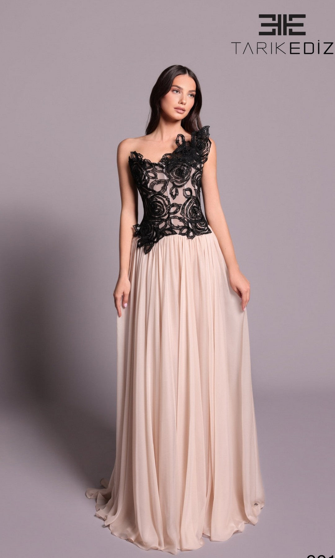 A woman poses against a plain light grey background wearing the Tarik Ediz 99101 long formal dress in pearl, featuring a chiffon A-line skirt and black lace-fitted bodice.